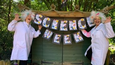 Two teachers dressed as white haired scientists standing either side of a banner that says 'Science Week'.