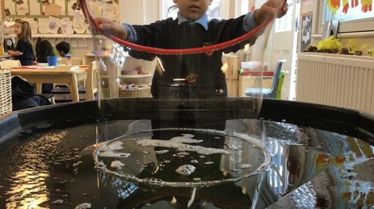 A child in a classroom making a giant bubble with a hula hoop.