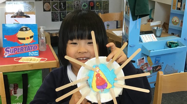 A school child holding up a piece of art they have made, a paper plate with a picture in the middle and lollipop sticks stuck around it.