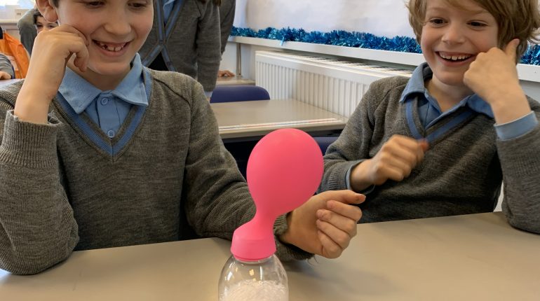 Two childrne in school uniforms looking at a bottle filled with white liquid, with a balloon stretched over the top.