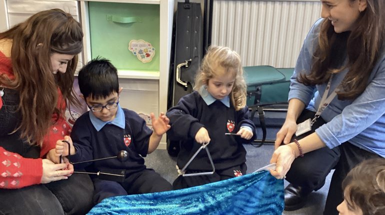 children playing with instruments as the teachers hold a cloak